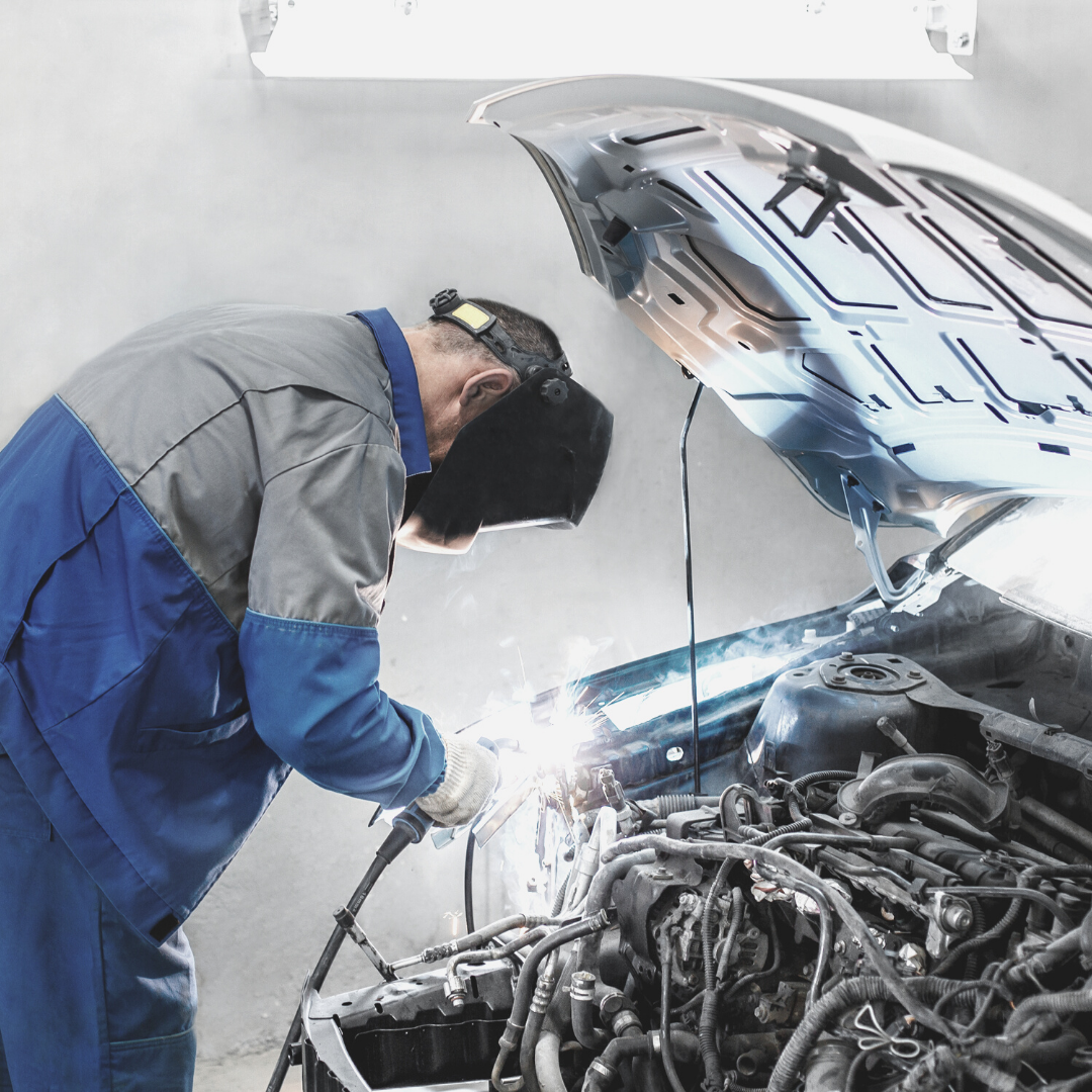 man in blue and grey jumpsuit wearing mask welding under automobile bonnet